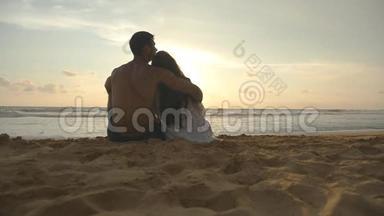 年轻的浪漫夫妇正在享受美丽的日落，坐在海滩上拥抱。 一个女人和一个男人坐在一起