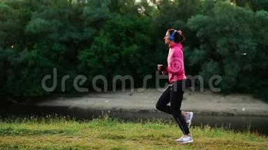 戴着长头发耳机的布鲁内特早上在公园里沿着河边跑步，夏天的日出时穿着粉红色的衣服