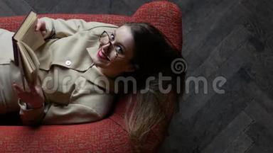 漂亮的年轻女子戴着米色的眼镜，坐在红色的沙发上看书，嬉戏地招手