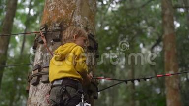 一个穿着安全装备的小男孩在森林探险公园的树梢上爬上了一条路线。 他是