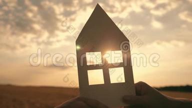 日落时分，在家中的田野里，双手紧握着纸模。 纸屋的剪影在光线下