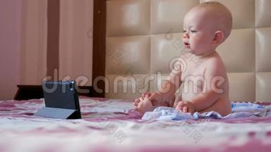 可爱的孩子坐在床上盯着平板电脑上的卡通。一岁的小婴儿，没有衣服
