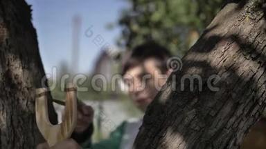 有趣的男孩躲在树后面，用弹弓瞄准户外