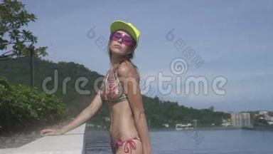 在阳光明媚的日子里，美丽的年轻女人穿着比基尼，黄色的帽子和紫色的太阳镜在无限的屋顶游泳池里摆姿势。