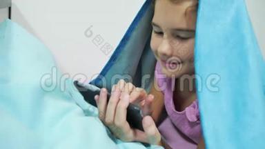 十几岁的女孩在毯子下<strong>玩</strong>智能<strong>手机</strong>。 在室内睡觉前的小女孩躺在<strong>床上上</strong>网