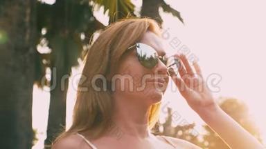 美丽时尚的金发女人戴着太阳镜，沿着棕榈树的小径散步。 手掌<strong>倒映</strong>在眼镜上.. 4K