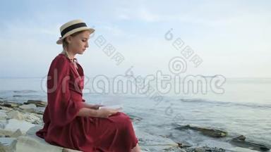 日落时分，一个漂亮的白人女孩穿着一件<strong>红色</strong>鲜艳的连衣裙和一顶草帽坐在<strong>海边</strong>的一块大石头上，看书