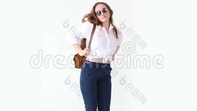 现代青年的时尚。 穿着牛仔裤、白衬衫、带皮背包和眼镜的时髦女孩在白色墙上摆姿势