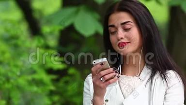 一位女士在公园里听着一部带耳机的智能手机里的音乐，一位美丽的女士用带耳机的智能手机听音乐