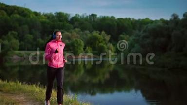 一个穿着粉红色夹克和黑色裤子的女孩戴着耳机在河边跑步，为马拉松做准备