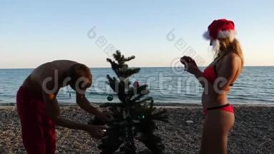 两个戴着圣诞帽的男人和女孩在海滩上<strong>迎接</strong>新年和圣诞节。 把圣诞树装饰在圣诞树上