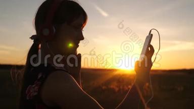 快乐的女孩在美丽的夕阳下，在智能手机上听音乐。 戴着耳机的年轻女孩