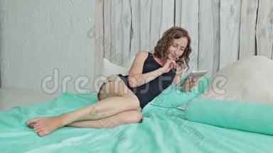 一位年轻的卷发女士躺在床上拿着平板电脑