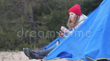 漂亮的女游客戴着红帽子坐在旅游帐篷里，喝着热水瓶里的茶