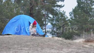 漂亮的女游客戴着红帽子坐在旅游帐篷里，喝着热水瓶里的茶