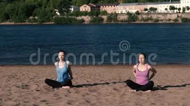 两个女人在<strong>城市</strong>河边的海滩上伸展瑜伽。 美丽的<strong>城市景色</strong>。 冥想姿势。
