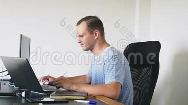 一个男人坐在家里的办公桌前，在家里的电脑和笔记本电脑里工作。