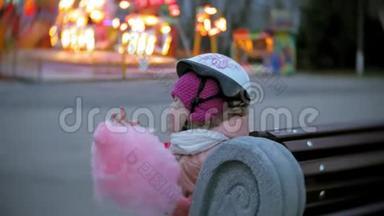 美丽的小女孩坐在<strong>游乐园</strong>的长凳上，吃着滚滑轮滑冰鞋上滚来滚去的粉红色甜棉糖