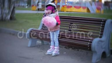美丽的小女孩坐在游乐园的长凳上，吃着滚滑轮滑冰鞋上滚来滚去的粉红色甜棉糖