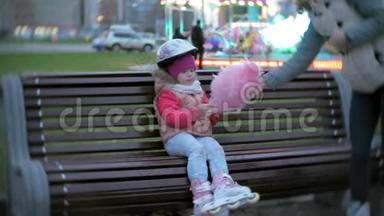 美丽的小女孩坐在游乐园的长凳上，吃着<strong>滚</strong>滑轮滑冰鞋上<strong>滚</strong>来<strong>滚</strong>去的粉红色甜棉糖