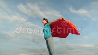 美丽的女孩超级英雄穿着红色斗篷站在田野上，斗篷随风飘扬。 慢动作。 女孩的梦想