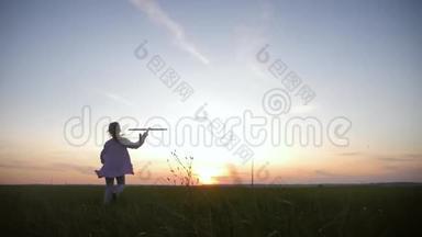 可爱的女孩在日落时在田里玩玩具木飞机。 儿童在木飞机上玩的剪影