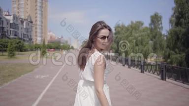 迷人的年轻女孩戴着太阳镜和一件白色的夏季时装长裙走在户外。 一个美丽的人