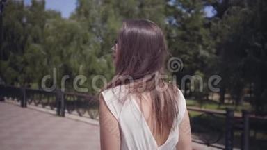 肖像迷人的年轻女孩戴着太阳镜和一件白色的夏季时装长裙走在户外。 休闲