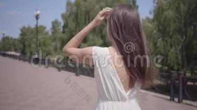 可爱的年轻女孩戴着太阳镜和一件白色的夏季时装长裙走在户外。 一个漂亮女人的闲情逸致