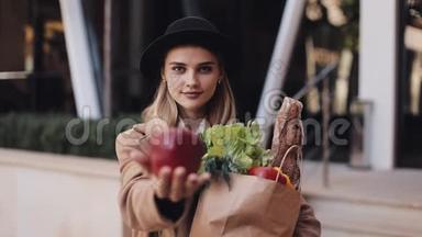 年轻漂亮的女人穿着时髦的外套站在街上，手里拿着一包产品，微笑着看着。