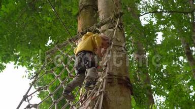 一个穿着安全装备的小男孩爬上森林探险公园的一条路线。 他爬在高绳上. 室外