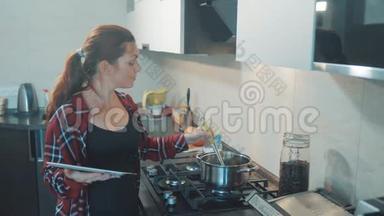 女人在厨房煮汤锅，防止锅里的水，看看数字平板上的食谱。 妇女