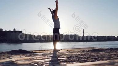 年轻的白种人通过在平静的河流附近的海滩上练习瑜伽健身运动放松身心，背景是城市