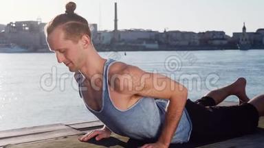 年轻的白种人通过在平静的河流附近的海滩上练习瑜伽健身运动放松身心，背景是城市