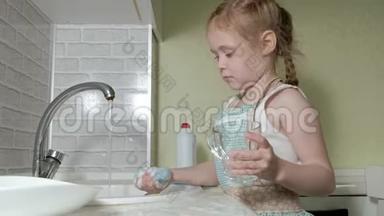 穿着<strong>围裙</strong>的漂亮小<strong>女孩</strong>站在椅子上，在明亮的厨房里，用海绵洗碗，帮助