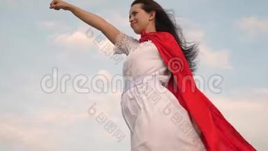 美丽的超级英雄女孩站在一片红色斗篷的田野上，斗篷在风中飘扬。 女孩梦想成为一个