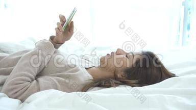 快乐的亚洲女人早上在床上使用智能手机。 亚洲女人在床上用智能手机检查社交应用程序。