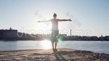 年轻的白种人通过在平静的河流附近的海滩上练习瑜伽健身运动放松<strong>身心</strong>，背景是城市