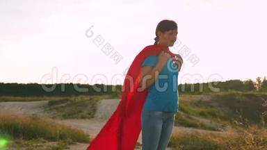 美丽的女孩超级英雄穿着红色<strong>斗篷</strong>站在田野上，<strong>斗篷</strong>随风飘扬。 慢动作。 一个年轻的女孩