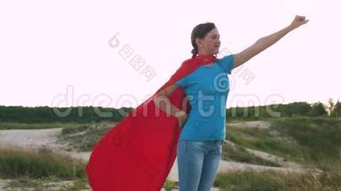 女孩梦想成为超级英雄。 美丽的女孩超级英雄穿着红色斗篷站在田野上，斗篷飘扬在田野上