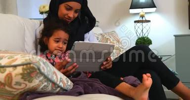 年轻的母亲在家里的沙发上和女儿一起使用数码平板电脑