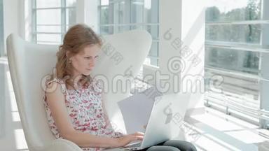 漂亮的女商人坐在玻璃办公室的白色椅子上，在笔记本电脑后面工作