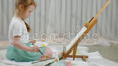 漂亮的红发女孩坐在画架前面的画室的地板上，画画笔和彩色颜料
