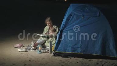 妈妈和她美丽的女儿晚上在海滩上的旅游帐篷附近读了一<strong>本书</strong>
