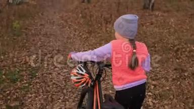一个高加索儿童在秋天公园骑自行车走路。 小女孩在森林里走着黑色的橙色循环。 孩子是干的