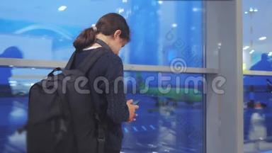 女儿旅行者带背包飞机航班机场概念。 带着智能手机的少女望着窗外
