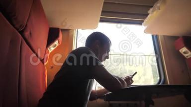 男子剪影旅行坐在火车车厢里，坐在窗边，一辆智能手机坐在铁路上戴着耳机