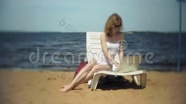 一个穿着白色比基尼的年轻女孩躺在沙滩上的躺椅上晒太阳，正在使用笔记本电脑