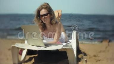 一个穿着白色比基尼的年轻女孩躺在<strong>沙滩上</strong>的躺椅<strong>上晒太阳</strong>，正在使用笔记本电脑
