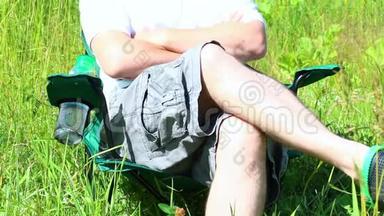一个人坐在一片灯火通明的草地上的野<strong>餐椅</strong>上。 他休息一下，关上一瓶水，把它放在一个支架上，然后把他的手